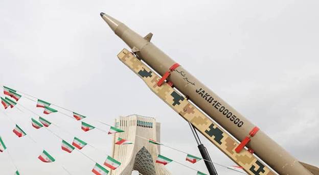 A missile displayed vertically with military camouflage patterns, set against a cloudy sky and featuring flags in the background, with a prominent building structure in the distance.