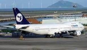 A large passenger airplane with a distinctive tail design parked at an airport, with wind turbines visible in the background.
