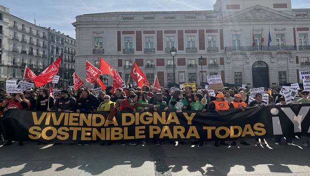 A large group of protesters holding a banner that reads 'Vivienda digna y sostenible para todas' (Dignified and sustainable housing for all) in front of a government building, with red flags and various signs.