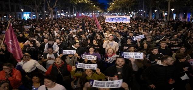 A large crowd of people protesting in the street, holding signs and banners. The scene is lively with individuals expressing their opinions, surrounded by trees and streetlights.