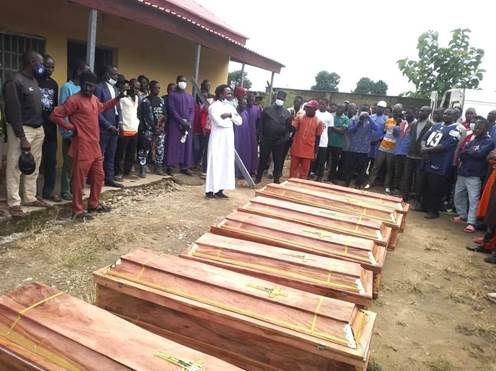 A gathering of mourners surrounding several wooden coffins, with a speaker addressing the crowd, in an outdoor setting.