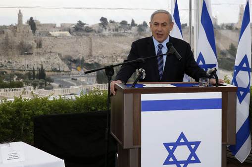 Man in a suit speaking at a podium with an Israeli flag, with Jerusalem's historic skyline in the background.