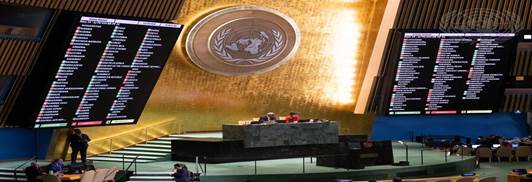 View of the United Nations General Assembly hall, featuring a large emblem on the wall and digital voting displays on either side.