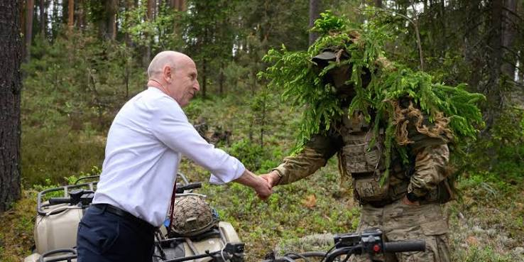 A civilian in a white shirt shakes hands with a military personnel camouflaged in foliage, set in a forested area with vegetation and all-terrain vehicles in the background.