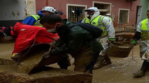 Rescue workers in protective gear assist with cleanup efforts in a muddy and flooded area, carrying buckets and working together in challenging conditions.