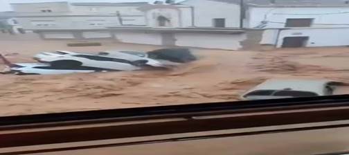 A flooded street with several cars partially submerged in muddy water, surrounded by buildings.