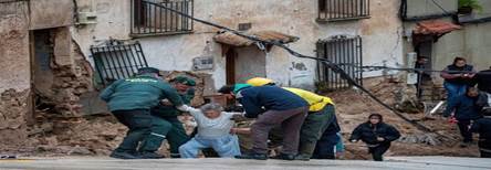 Rescue workers assisting an elderly person near a damaged building with collapsed walls and exposed debris.